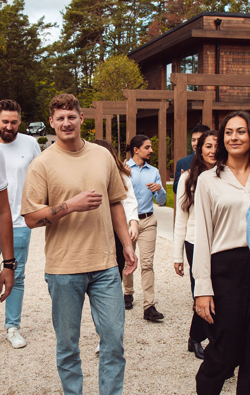 A group of young adults walking together in a friendly, relaxed manner. A man in a beige T-shirt and blue jeans is at the forefront, with a visible tattoo on his left arm. Behind him, various other individuals in smart-casual attire converse and enjoy the walk. They are in an outdoor setting with a wooden structure and green trees in the background, which could indicate a corporate retreat or team-building environment.