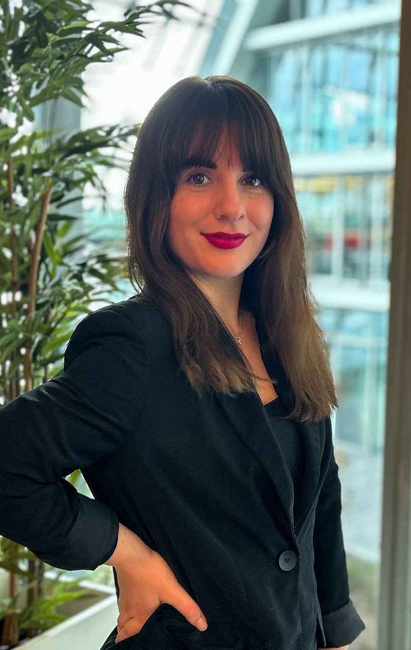 A portrait of a young woman with shoulder-length brunette hair and bangs, wearing a black blazer. She has a subtle smile and is wearing red lipstick. In the background, there are indoor plants and a bright, airy atrium with a glass facade, suggesting a modern office environment.