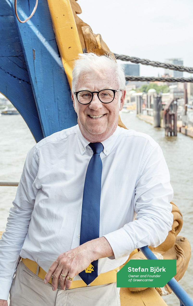 Portrait of Stefan Björk, a smiling elderly gentleman with white hair and glasses, dressed in a light blue shirt with rolled-up sleeves, a dark blue tie, and beige trousers with a yellow belt. He is leaning casually against a large blue and yellow industrial object, possibly part of a ship, with a harbor scene blurred in the background. A caption identifies him as the Owner and Founder of Greencarrier.