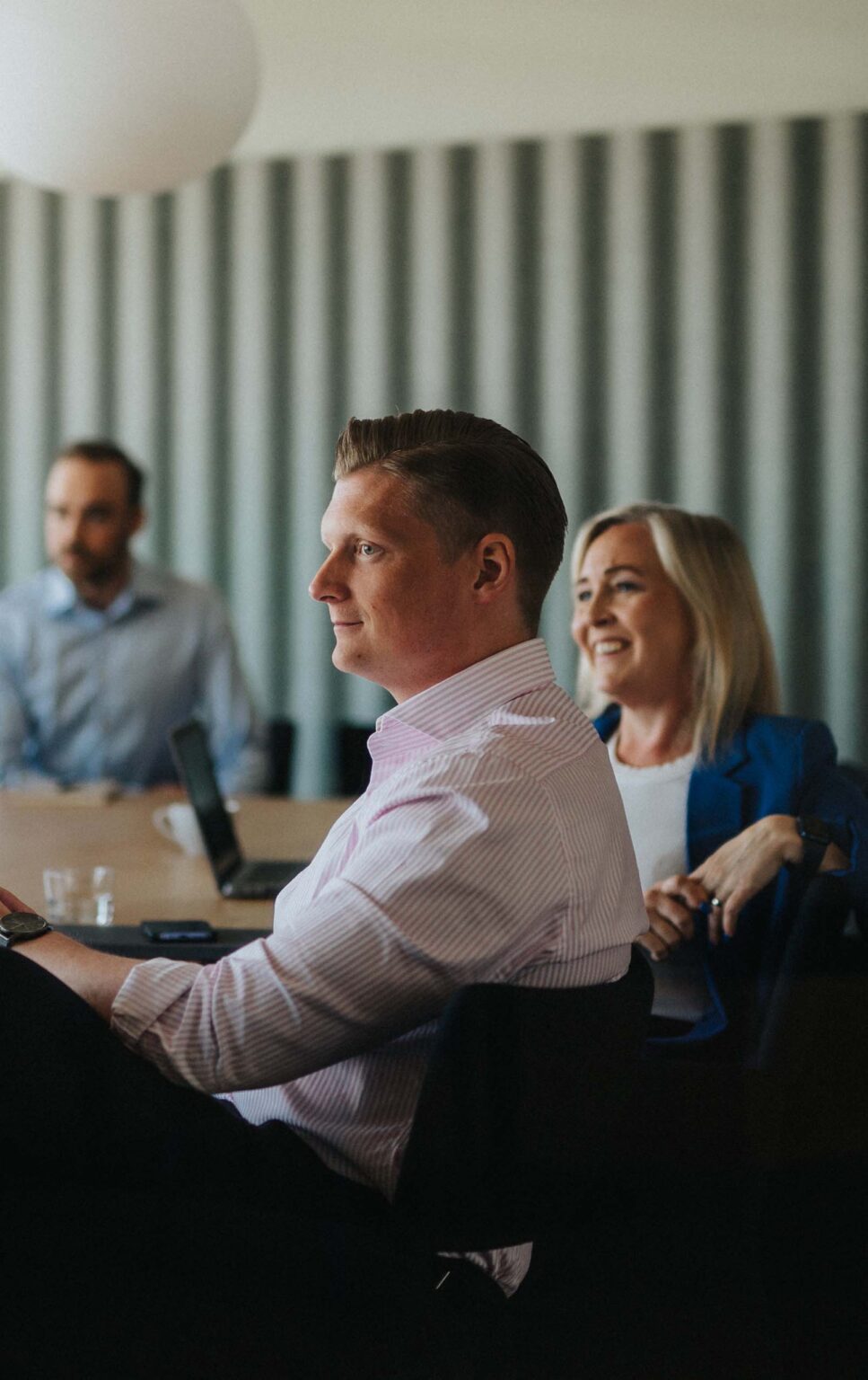 A group of people is sitting at a conference table during a meeting. The focus is on a man in a light pink striped shirt, who is seated in profile and looking attentively forward. Next to him, a woman with blonde hair and a blue blazer is smiling. In the background, another man, slightly out of focus, is also seated and appears to be engaged in the discussion. A laptop and a coffee cup are visible on the table. The setting is professional, with a modern, softly lit interior featuring vertical blinds.