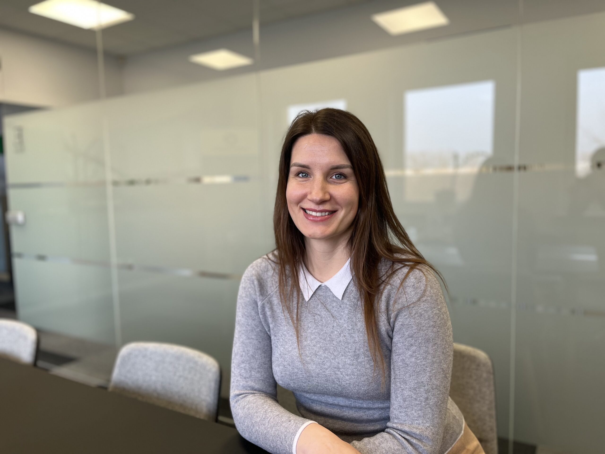 A woman with long brown hair and a friendly smile is seated at a conference table in a modern office setting. She is wearing a gray sweater over a white collared shirt. Behind her, there is a frosted glass wall with geometric patterns, allowing light to filter through while maintaining privacy. The atmosphere is professional and welcoming.