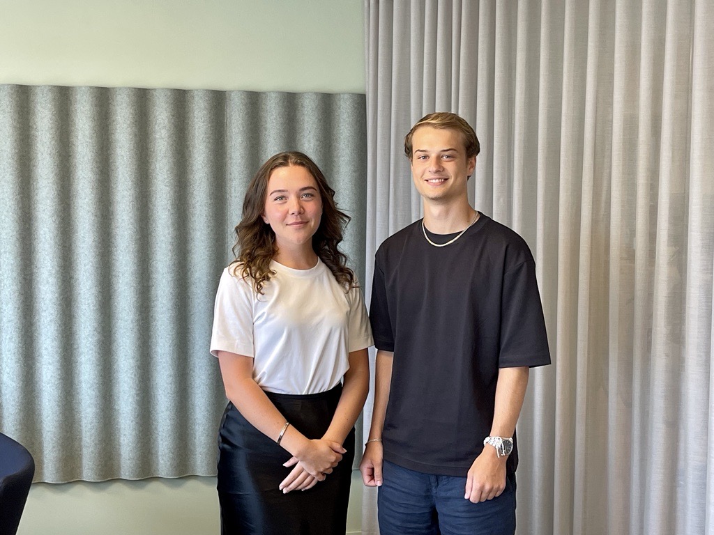 Two young people are standing side by side in an office environment. The woman on the left has long wavy brown hair and is wearing a white t-shirt paired with a black skirt. The man on the right has short light brown hair and is dressed in a black t-shirt with a chain necklace. Both are smiling softly, creating a warm and approachable atmosphere. The background features soundproof panels and curtains, suggesting a well-designed office space focused on comfort and functionality.