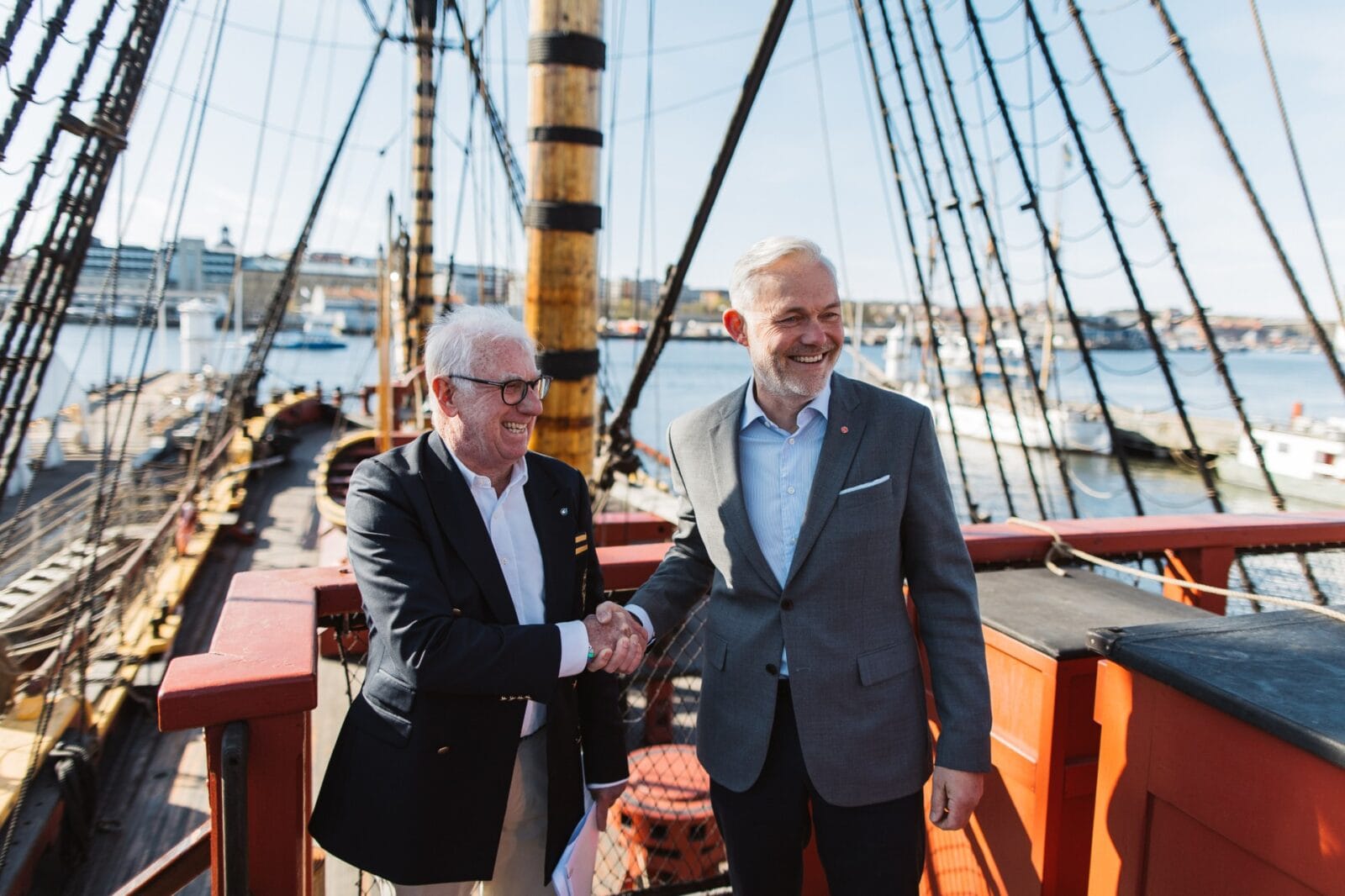 Two men in suits shake hands on the deck of a historic sailing ship with rigging and masts in the background