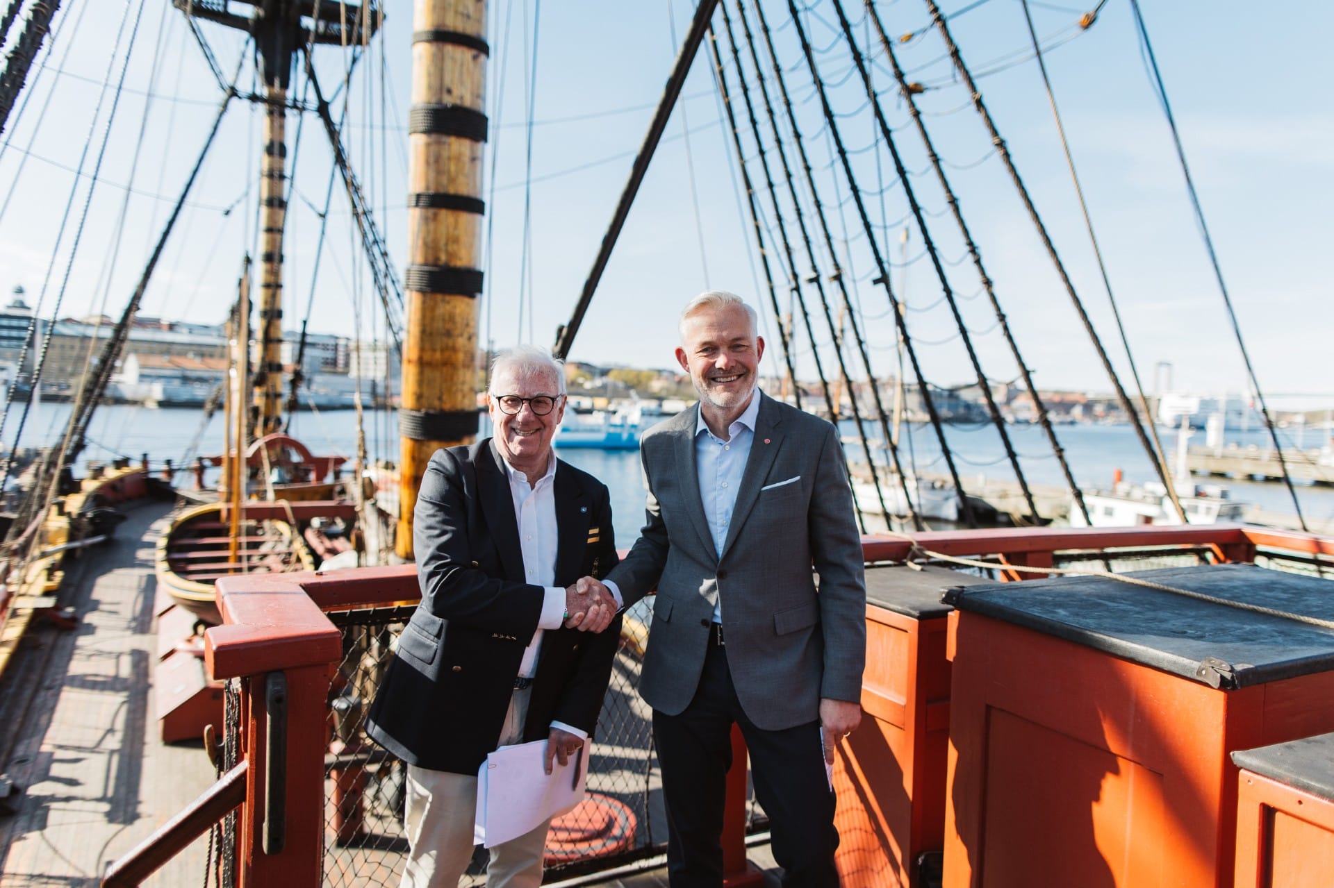 Two businessmen in suits shake hands on a ships deck with tall rigging and masts in the background