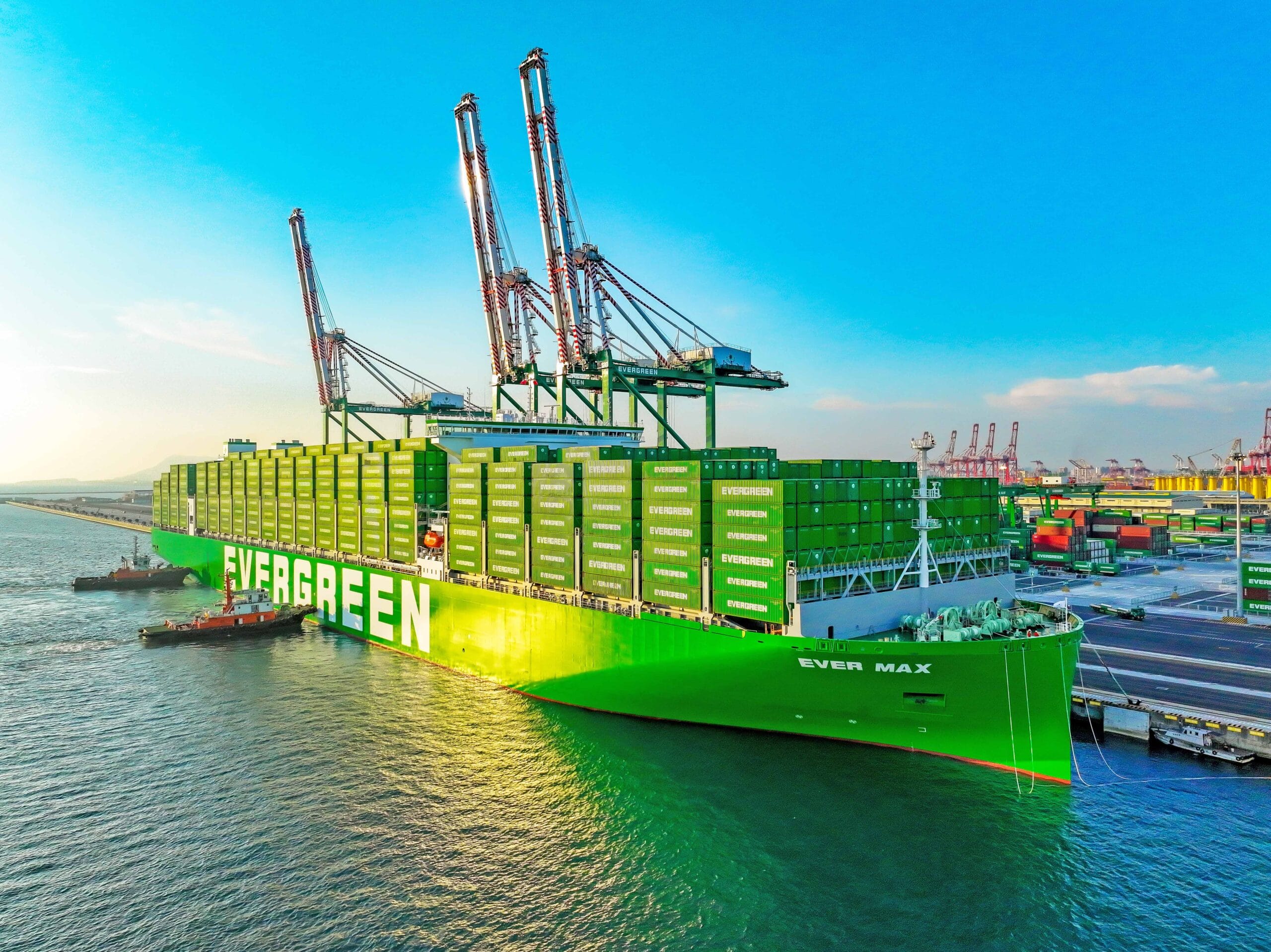 Green cargo ship loaded with stacked shipping containers labeled EVERGREEN at a busy port with tall cranes in the background