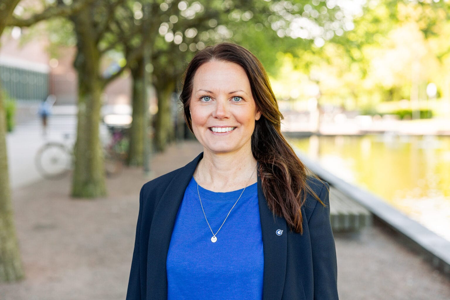 Woman with brown hair looking in to the camera and smile She wears a blue shirt and dark colored blazer Behind her is trees and a fontain in the background